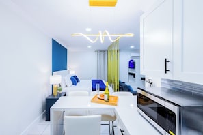 Et moderne køkken med indbygget ovn og mikroovn, et spiseområde med et hvidt bord og stole, samt en opholdsstue med sofa og blå puder.