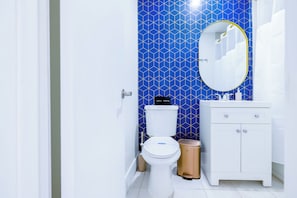Et badeværelse med et hvidt toilet, en håndvask med spejl og en skraldespand.