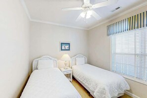 Ein Schlafzimmer mit zwei Betten, einem Deckenventilator, einem Nachttisch mit Lampe, einem gerahmten Bild an der Wand und einem Fenster mit Jalousien.
