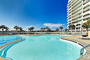 Uma piscina com design curvo, cercada por uma grade, ao lado de um hotel com vários andares e varandas.