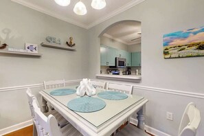 A dining area with a glass table, white chairs, and a view into the kitchen. - The Sandy Oyster Retreat (North Myrtle Beach)