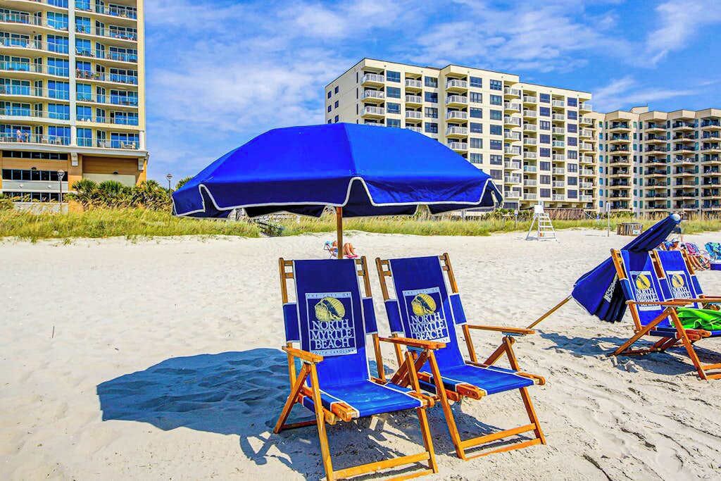 Cadirens de platja amb el logotip de North Myrtle Beach, un para-sol blau i un edifici al fons.