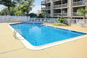 A rectangular swimming pool with a metal ladder on one side, surrounded by a white fence with signs, and a building with balconies in the background. - Shipwatch Pointe II L-302 (Myrtle Beach)