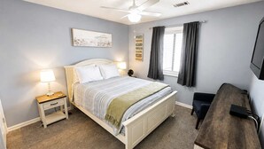 Ein Schlafzimmer mit einem Bett, Nachttischen, einem Deckenventilator, einem Fenster mit Vorhängen und einem Fernseher auf einem Holzboden.