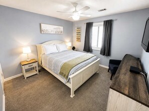 Ein Schlafzimmer mit einem Bett, Nachttischen, einem Deckenventilator, einem Fenster mit Vorhängen und einem Fernseher auf einem Holzboden.