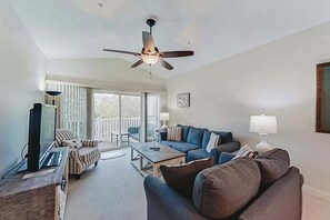 Ein Wohnzimmer mit Deckenventilator, Fernseher, Sofa, Couchtisch und Blick ins Freie durch eine Schiebetür.
