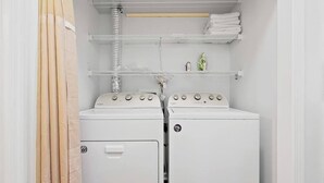Una sala de bugaderia amb dues piles de rentadores i assecadores, una prestatgeria de metall amb tovalloles plegades i una sortida d'aire per sobre de les assecadores.