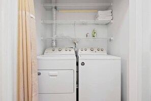 A laundry room with two stacked washers and dryers, a metal rack with folded towels, and a vent above the dryers. - Village at the Glens L-205 (Little River)