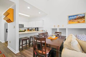 Een moderne keuken met witte kastjes, een eethoek met een houten tafel en stoelen, en een schilderij aan de muur.