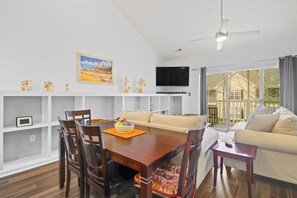 A modern living room with a dining area, a flat-screen TV mounted on the wall, and a large window offering a view of the outdoors. - Village at the Glens L-205 (Little River)