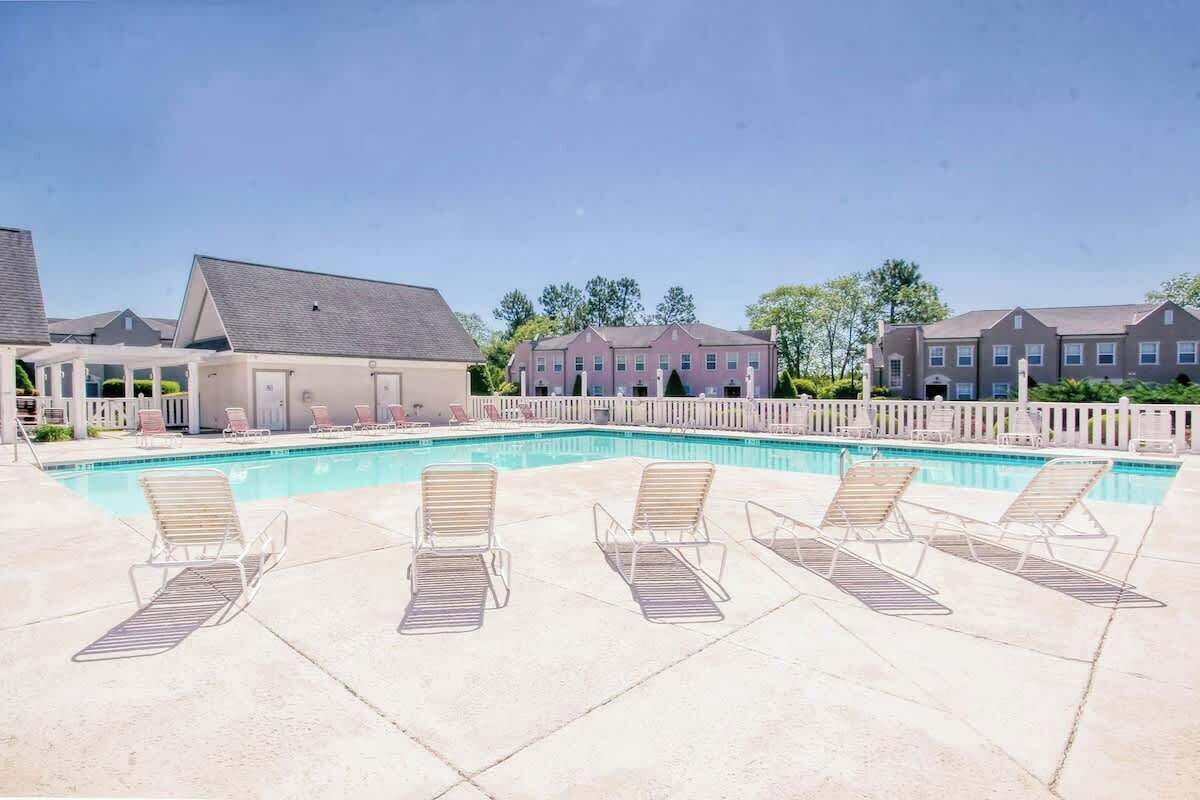 A swimming pool with lounge chairs, a covered area, and a row of houses in the background.