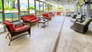 A covered outdoor seating area with wicker furniture and red cushions, large windows, and a view of a body of water. - Homey Beach Hideaway C (Myrtle Beach)