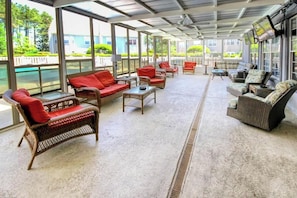 A covered outdoor seating area with wicker furniture and red cushions, large windows, and a view of a body of water. - Homey Beach Hideaway C (Myrtle Beach)