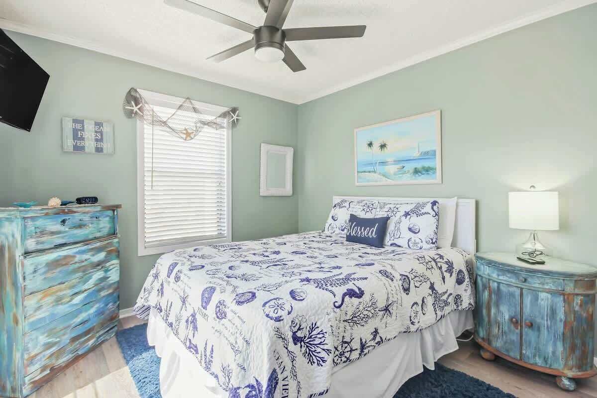 Une chambre à coucher comprenant un lit, une commode, une table de chevet, un ventilateur de plafond, une fenêtre avec des stores et un tableau accroché au mur.