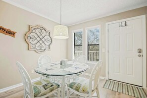 A dining area with a round glass table, white chairs, a hanging light fixture, and a door with a window view. - Hillside Unit 304 Condo (North Myrtle Beach)