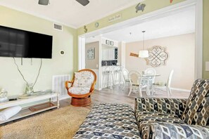 A living room with a sofa, a wicker chair, a flat-screen TV, and a dining area with white furniture.
