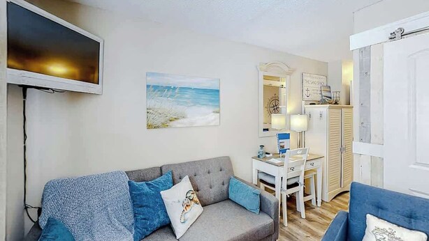 Uma sala de estar com um sofá cinzento, uma poltrona azul, uma televisão de tela plana, um quadro de uma praia e uma escrivaninha com uma luminária.