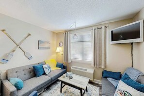 A living room with a sofa, a coffee table, a television, and a window with curtains. - The Sand Shack - Litchfield Inn 251 (Pawleys Island)