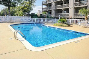 Ein rechteckiger Swimmingpool mit einer Metallleiter an der linken Seite, umgeben von einem weißen Zaun und einem Gebäude mit Balkonen im Hintergrund.