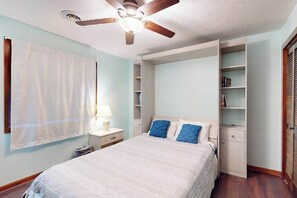 A bedroom with a bed, a ceiling fan, a nightstand, a lamp, a window with a curtain, and built-in shelving. - Shipwatch Pointe II Q-305 (Myrtle Beach)