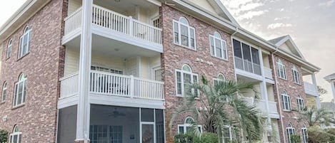 Un immeuble résidentiel à plusieurs étages, doté d’un balcon au rez-de-chaussée, d’un patio couvert et d’une façade en briques.