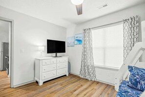 Ein Zimmer mit weißem Schminktisch, einem Flachbildfernseher, einem Fenster mit gemusterten Vorhängen und einem Bett mit einem Kissen mit Muster.