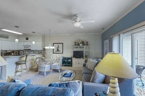 A living room with a sofa, armchairs, a coffee table, and a TV. There is a kitchen area with cabinets and a refrigerator. A ceiling fan is visible, and the room has a window. - Scotch Broom Unit E-104 (Little River)