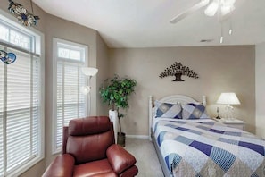 A bedroom with a bed, a chair, a nightstand, a lamp, a wall decoration, and a ceiling fan. - Scotch Broom Unit E-104 (Little River)