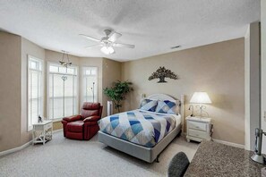A bedroom with a bed, a chair, a nightstand, a lamp, a ceiling fan, and a window with blinds. - Scotch Broom Unit E-104 (Little River)