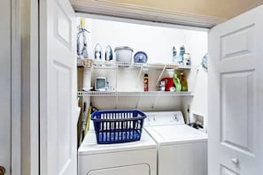 A laundry room with a washing machine, shelves with various items, and a blue basket. - Scotch Broom Unit E-104 (Little River)