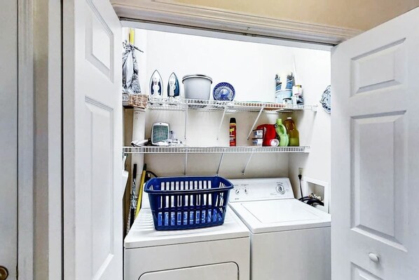 A laundry room with a washing machine, shelves with various items, and a blue basket. - Scotch Broom Unit E-104 (Little River)