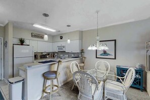 A kitchen with white cabinetry, a central island, bar stools, and wicker chairs. - Scotch Broom Unit E-104 (Little River)