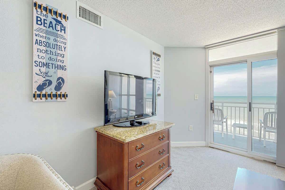 Ein Zimmer mit Fernseher auf einer Holzkassette, einem großen Fenster mit Blick auf den Strand und einem Poster an der Wand.
