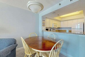 A dining area with a wooden table and chairs, a kitchen with white cabinets, and a ceiling light fixture. - Carolinian Beach Resort 0730 (Myrtle Beach)