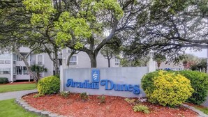 A sign for Arendian Dunes with a landscaped garden and a large tree in the foreground. - Arcadian Dunes 18-370 (Myrtle Beach)
