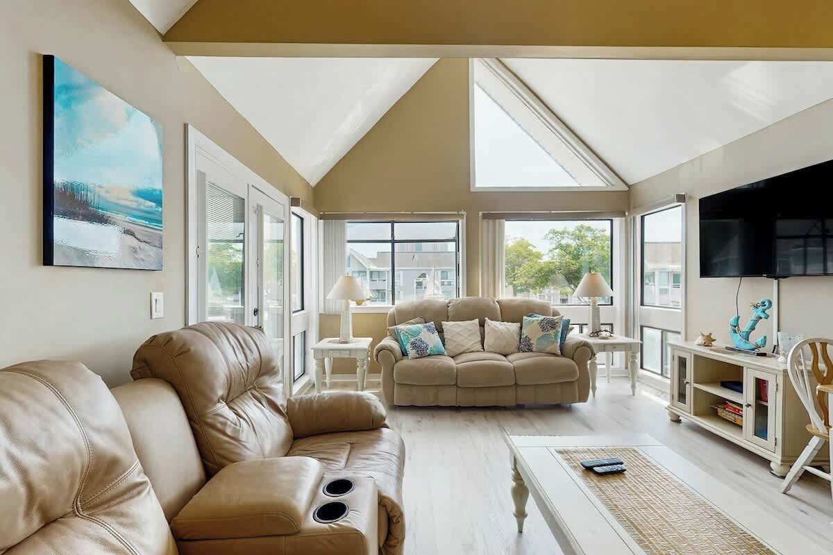 A spacious living room with a high ceiling, large windows, and a television.