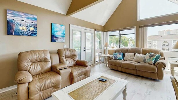 A spacious living room with two leather sofas, a coffee table, and large windows offering a view of the outside. - Arcadian Dunes 18-370 (Myrtle Beach)