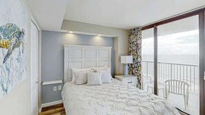 A bedroom with a bed, a nightstand, a window with a view, and a balcony with chairs. - Sands Ocean Club 1629 (Myrtle Beach)
