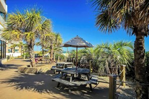 An outdoor seating area with wooden tables and benches, surrounded by palm trees and a clear blue sky. - Sands Ocean Club 1629 (Myrtle Beach)