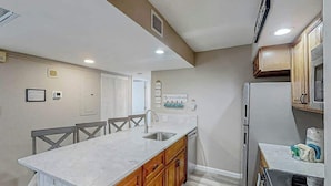 A kitchen with a marble countertop, wooden cabinets, a stainless steel sink, and a refrigerator. - Sands Ocean Club 1629 (Myrtle Beach)