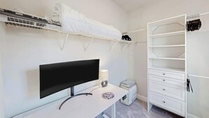 Et soveværelse med seng, skrivebord, fjernsyn, garderobe og en reol med tøj.
