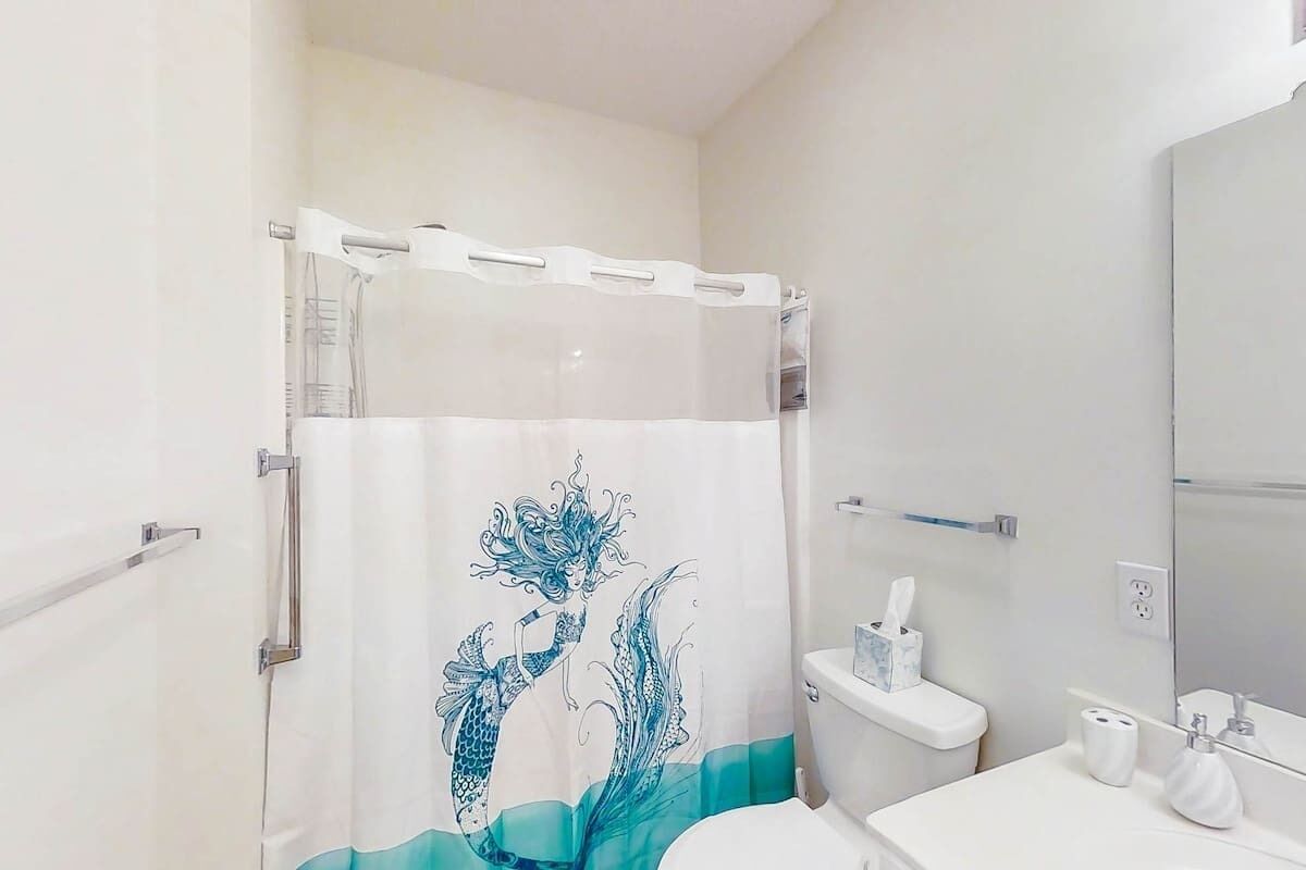 Uma casa de banho com cortina de chuveiro com desenho de sereia, sanita, lavatório com espelho e porta-toalhas.