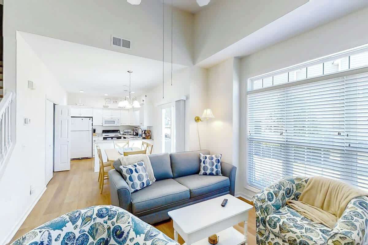A modern living space with a sofa, a coffee table, and a patterned armchair. A kitchen with white cabinets and a dining area is visible in the background.