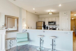 A modern kitchen with white cabinets, a stainless steel refrigerator, a microwave, and a coffee maker. There are two bar stools and a light blue chair. - Waterway Landing 205-H (North Myrtle Beach)