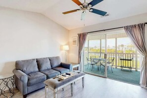 Ein Zimmer mit grauem Sofa, einem kleinen Tisch, einem Deckenventilator und Blick auf einen Außenbalkon mit Tisch und Stühlen.