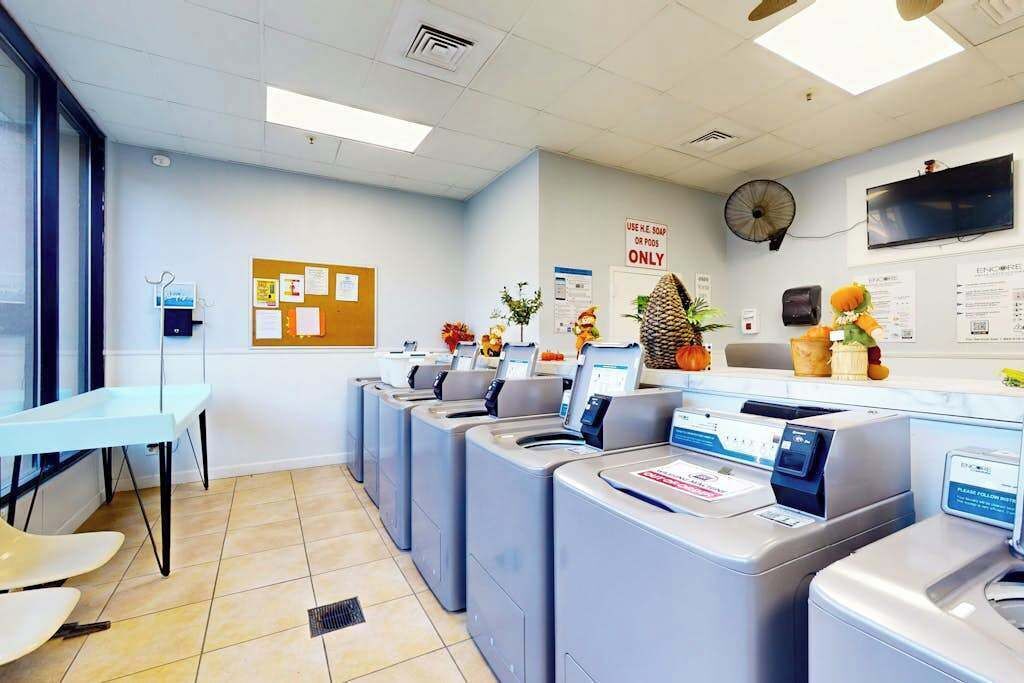A laundry facility with multiple washing machines, a table with chairs, and a wall-mounted fan.