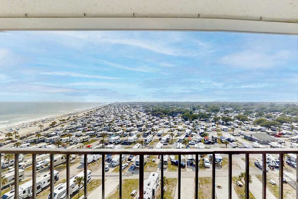 Un grand terrain de camping pour véhicules récréatifs, avec de nombreux véhicules stationnés en rangées bien ordonnées, une plage en arrière-plan et un ciel dégagé.