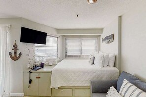 Une chambre à coucher comprenant un lit, un téléviseur fixé au mur, un bureau avec un ordinateur et une fenêtre avec des stores.