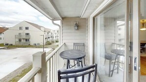 A balcony with a table and chairs, a view of neighboring buildings, and a glass door leading to an interior space. - Greystone Condos 203 (Myrtle Beach)