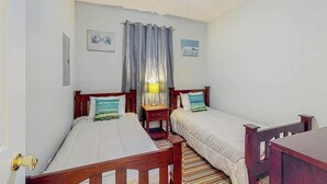 Ein Zimmer mit zwei Einzelbetten, einem Deckenventilator, einem Fenster mit Vorhängen und einem kleinen Tisch mit einer Lampe.